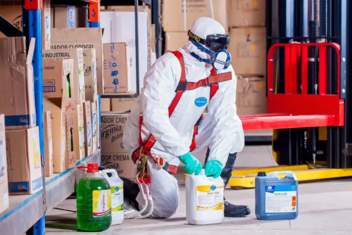 A technician in protective equipment handling industrial chemical containers in a warehouse.